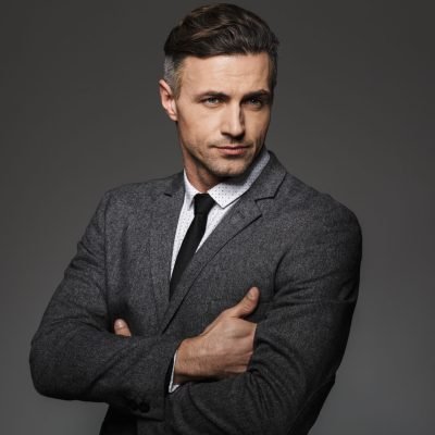 Portrait of masculine man wearing business suit posing on camera Portrait of masculine man wearing business suit posing on camera with serious look keeping arms crossed isolated over gray background